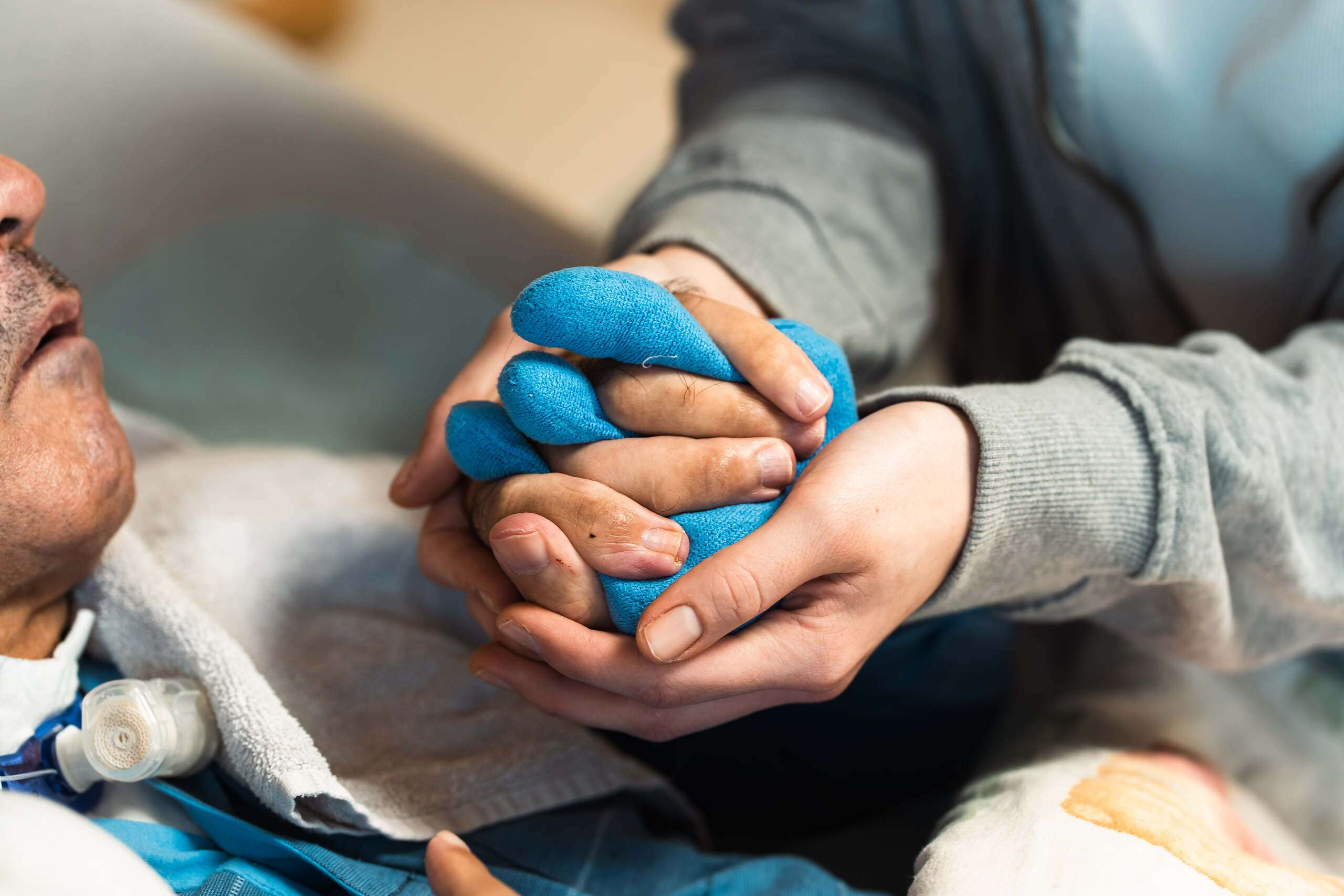 Eine Person hält die Hände eines anderen mit blauer Bandage während der Pflege in einem medizinischen Umfeld.